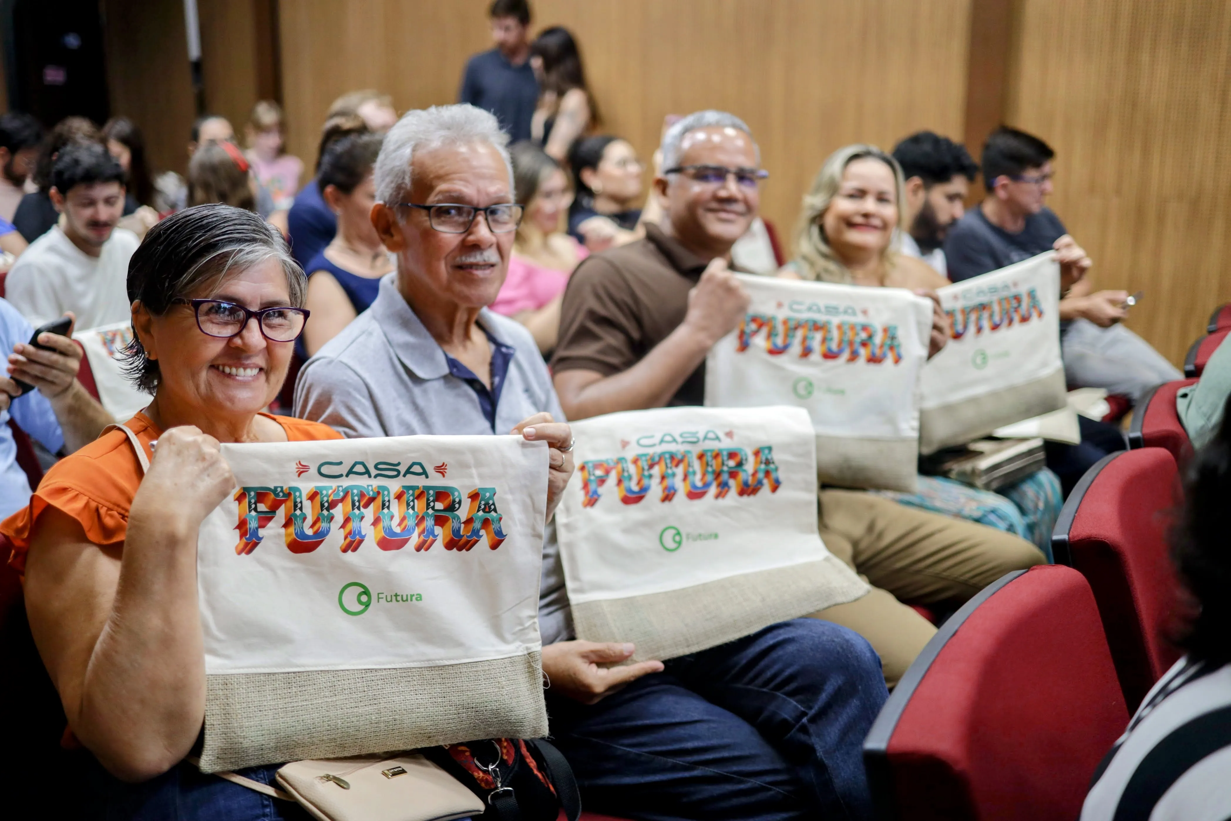 Uma sala iluminada com pessoas sentadas em cadeiras vermelhas. Na frente, um grupo de quatro pessoas mostra bolsas com uma estampa colorida. No desenho está escrito Casa Futura.