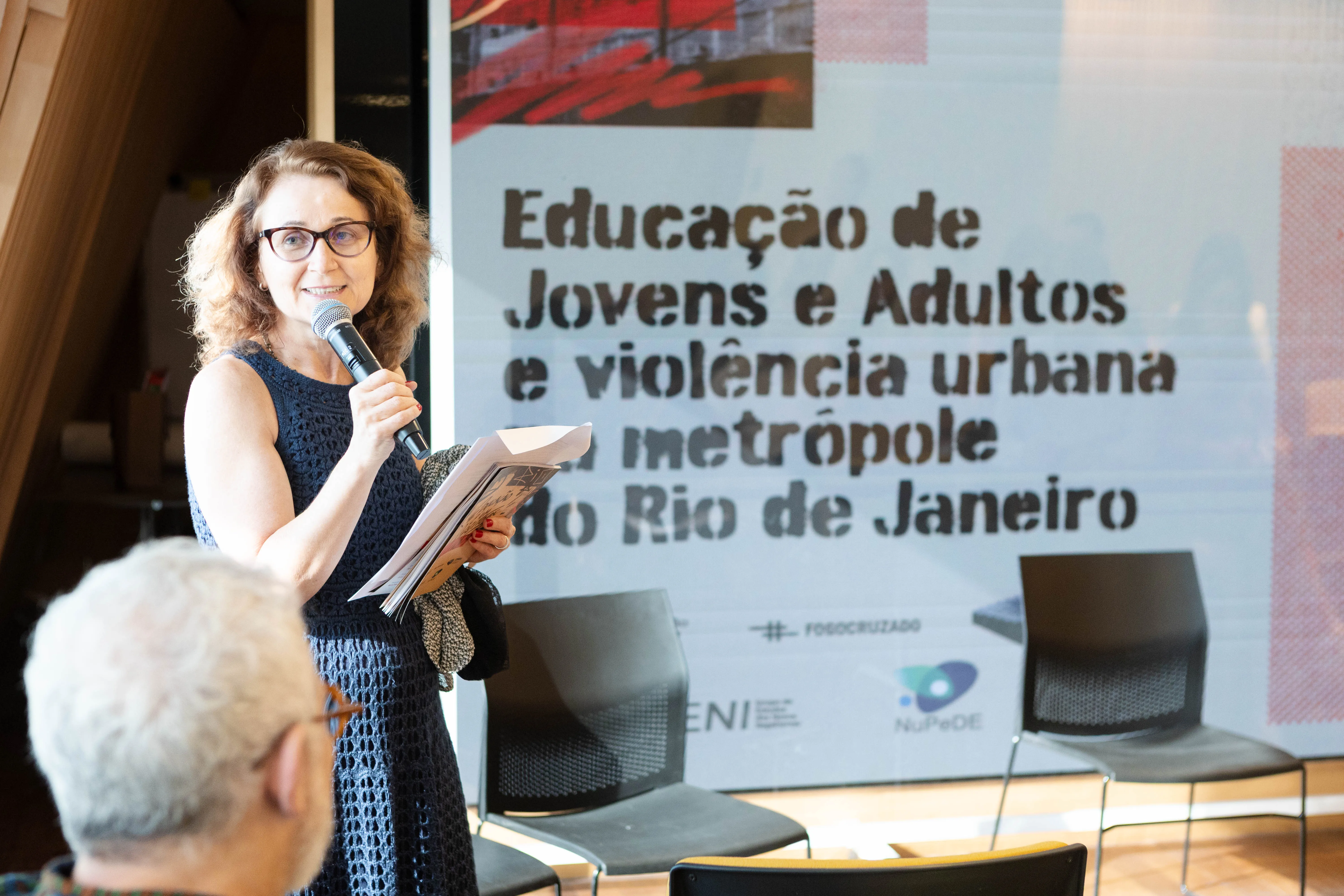 A palestrante segura um microfone e uma publicação enquanto fala ao público. Atrás dela, um telão exibe o título “Educação de Jovens e Adultos e violência urbana na metrópole do Rio de Janeiro”. A cena ocorre em um ambiente de auditório, com cadeiras organizadas ao fundo.
