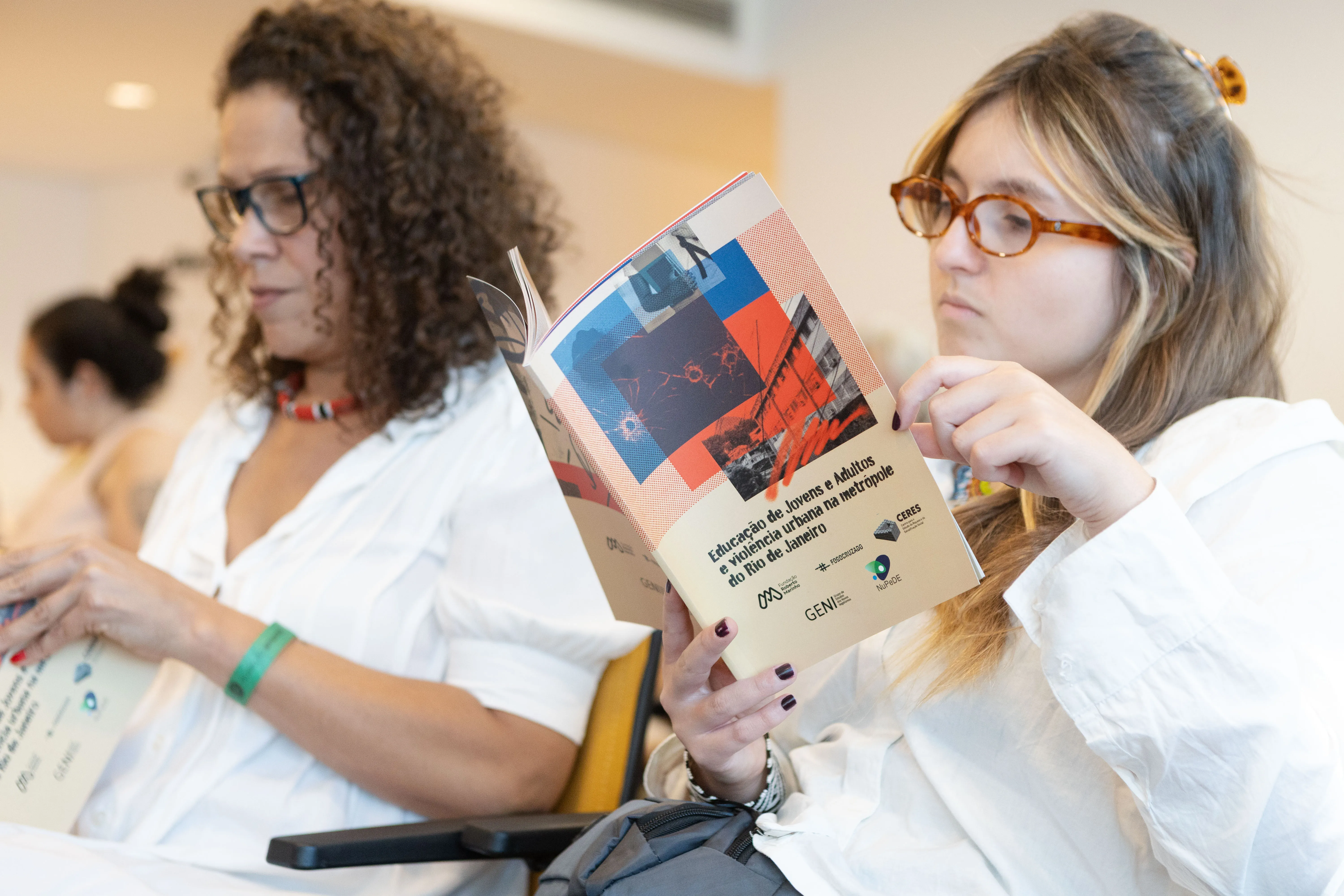 Participantes do evento acompanham a leitura da publicação da pesquisa “Educação de Jovens e Adultos e violência urbana na metrópole do Rio de Janeiro”. Elas estão sentadas no auditório, concentradas no material distribuído.