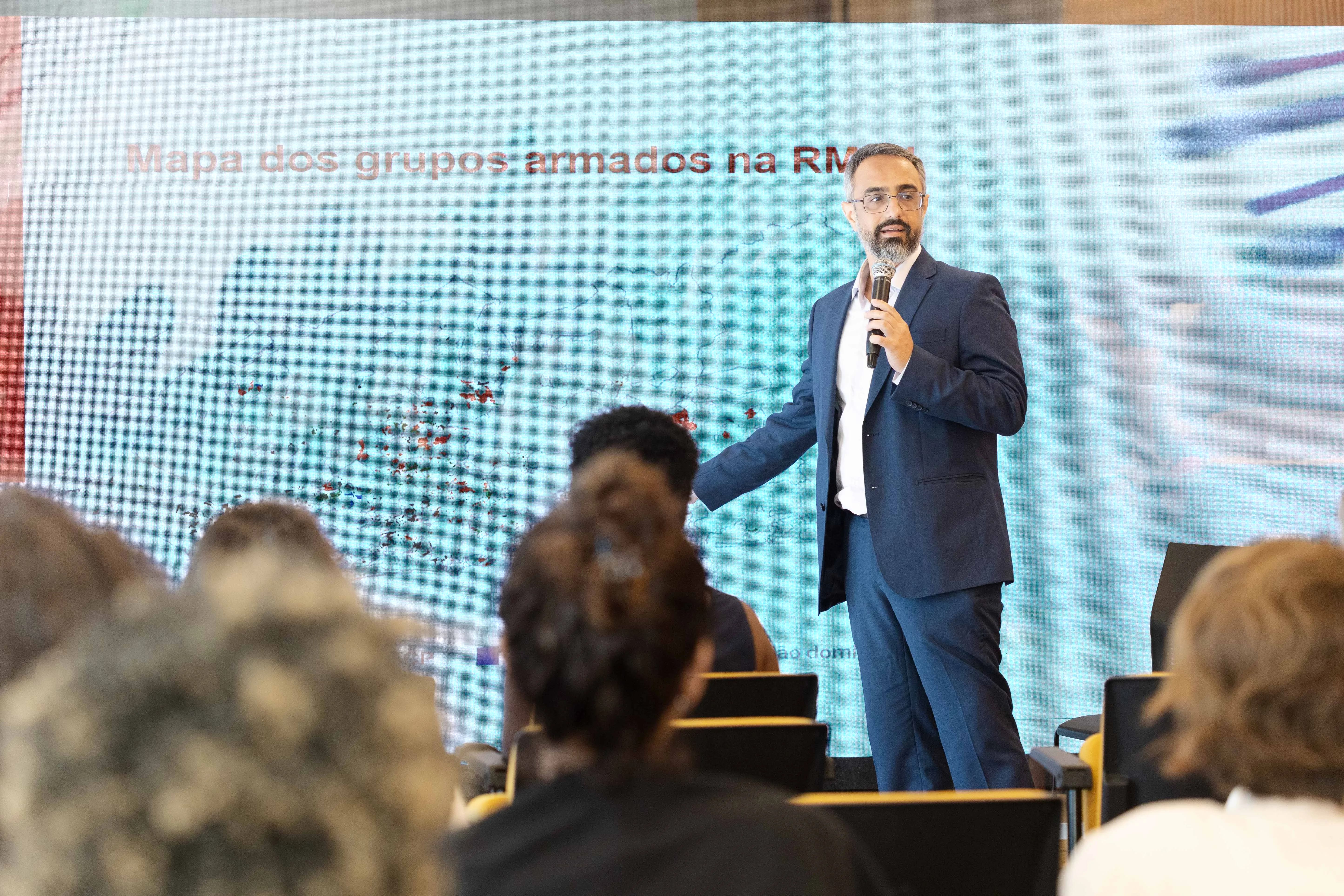 Um pesquisador aponta para um mapa exibido no telão atrás dele, identificado como “Mapa dos grupos armados na RMRJ”. Ele segura um microfone e dialoga com a plateia, que aparece desfocada no primeiro plano.