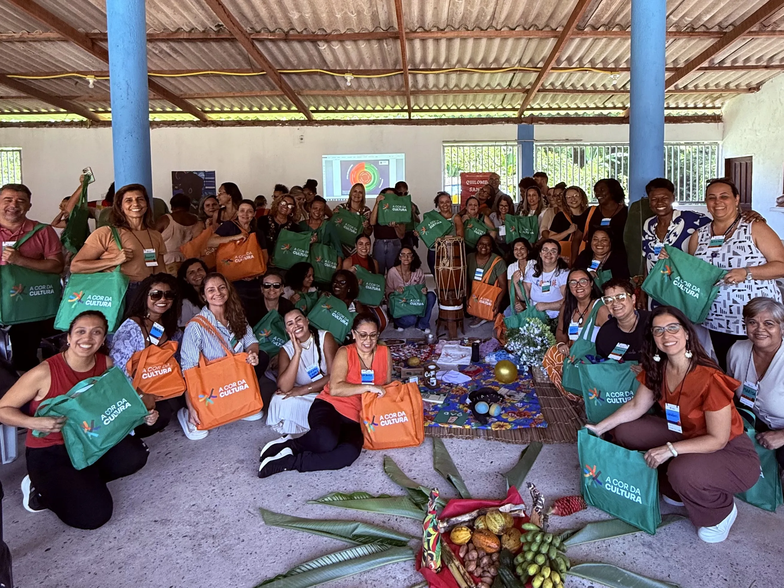 o grupo posa junto, sorridente, segurando bolsas com a identidade visual de “A Cor da Cultura”, indicando a conclusão de uma atividade, oficina ou formação. No centro, elementos como frutas, folhas e objetos simbólicos sugerem uma prática ritualística ou pedagógica conectada à cultura e à ancestralidade.