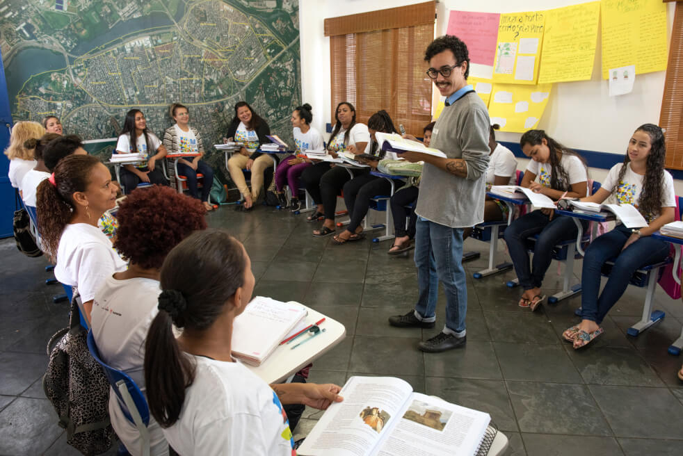 Alunos adolescentes em volta do professor, enquanto ele lê um livro para elas