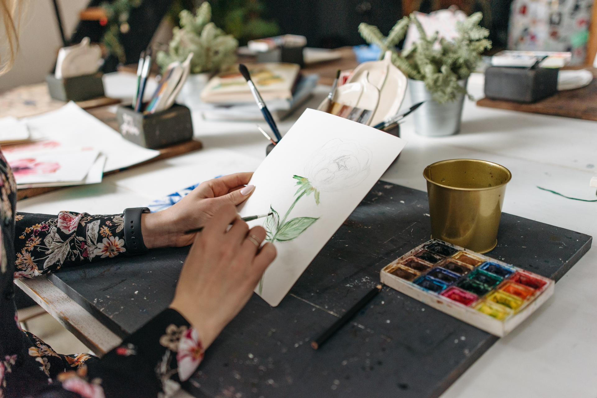 Na imagem, mãos de uma mulher pintam uma planta verde em um papel em branco. Ao lado, uma pequena lata e potes com as tintas coloridas.