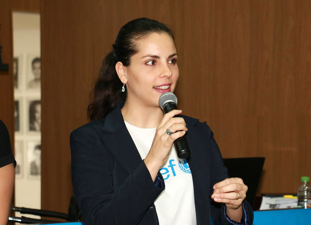Uma mulher branca, de cabelos escuros presos em um rabo de cavalo, fala com um microfone na mão.