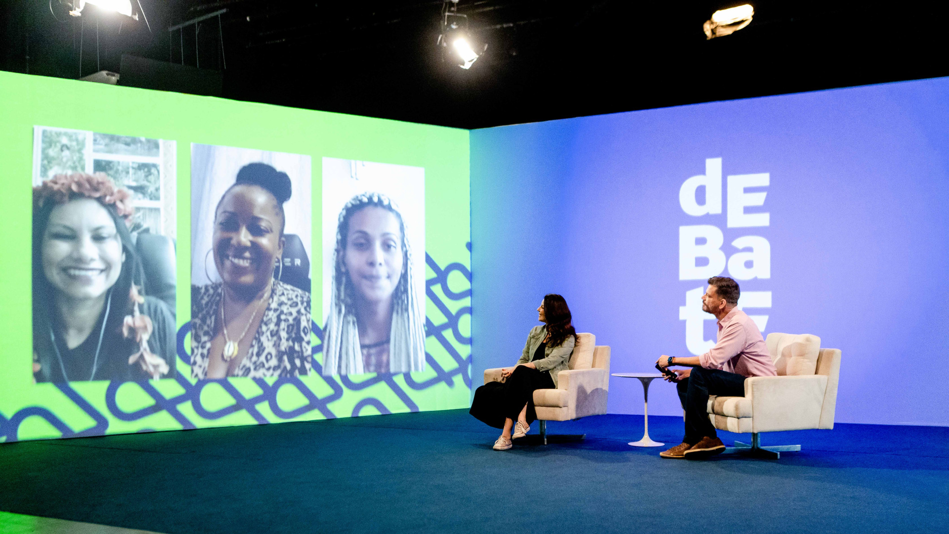 Três mulheres, à esquerda, estão com suas imagens projetadas enquanto conversam por videoconferência com duas pessoas, uma mulher e um homem, que estão sentados, à direita.