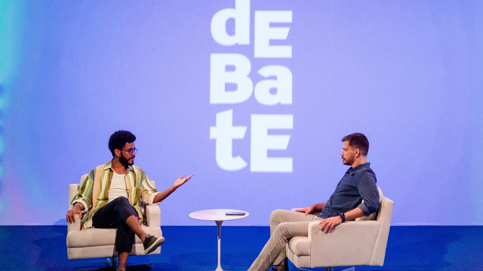 Dois homens estão sentados em poltronas, um de frente para o outro.