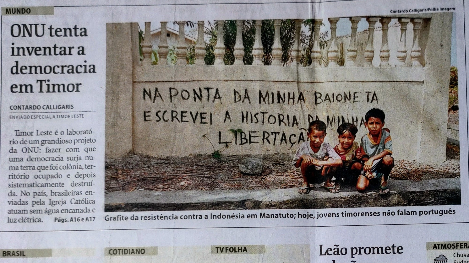 Foto de folha de jornal com matéria "ONU tenta inventar a democracia em Timor". Ao lado do título, a imagem de três crianças agachadas diante da seguinte frase, escrita no muro: "Na ponta da minha baioneta, escreveis a história da minha libertação".
