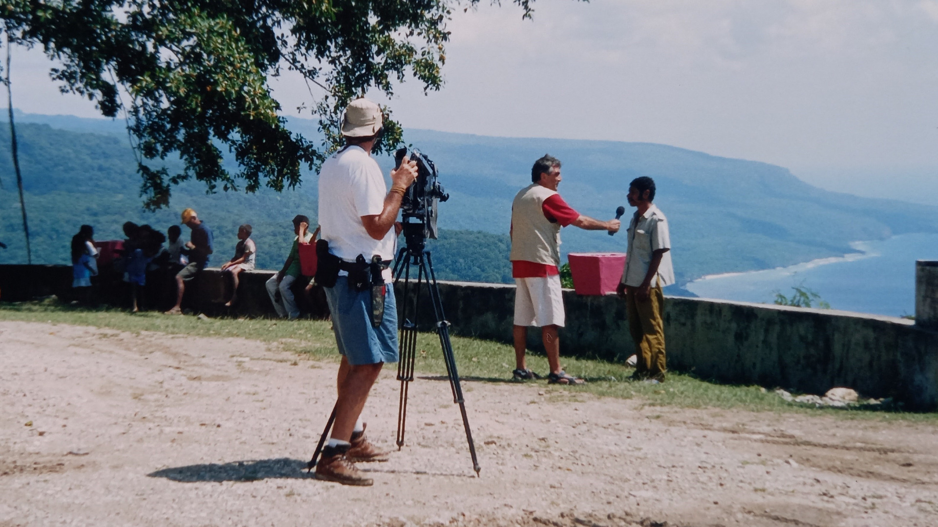 Em um mirante, um homem aparece em primeiro plano, filmando um repórter que entrevista outro homem. Ao fundo, abaixo, montanha e mar.