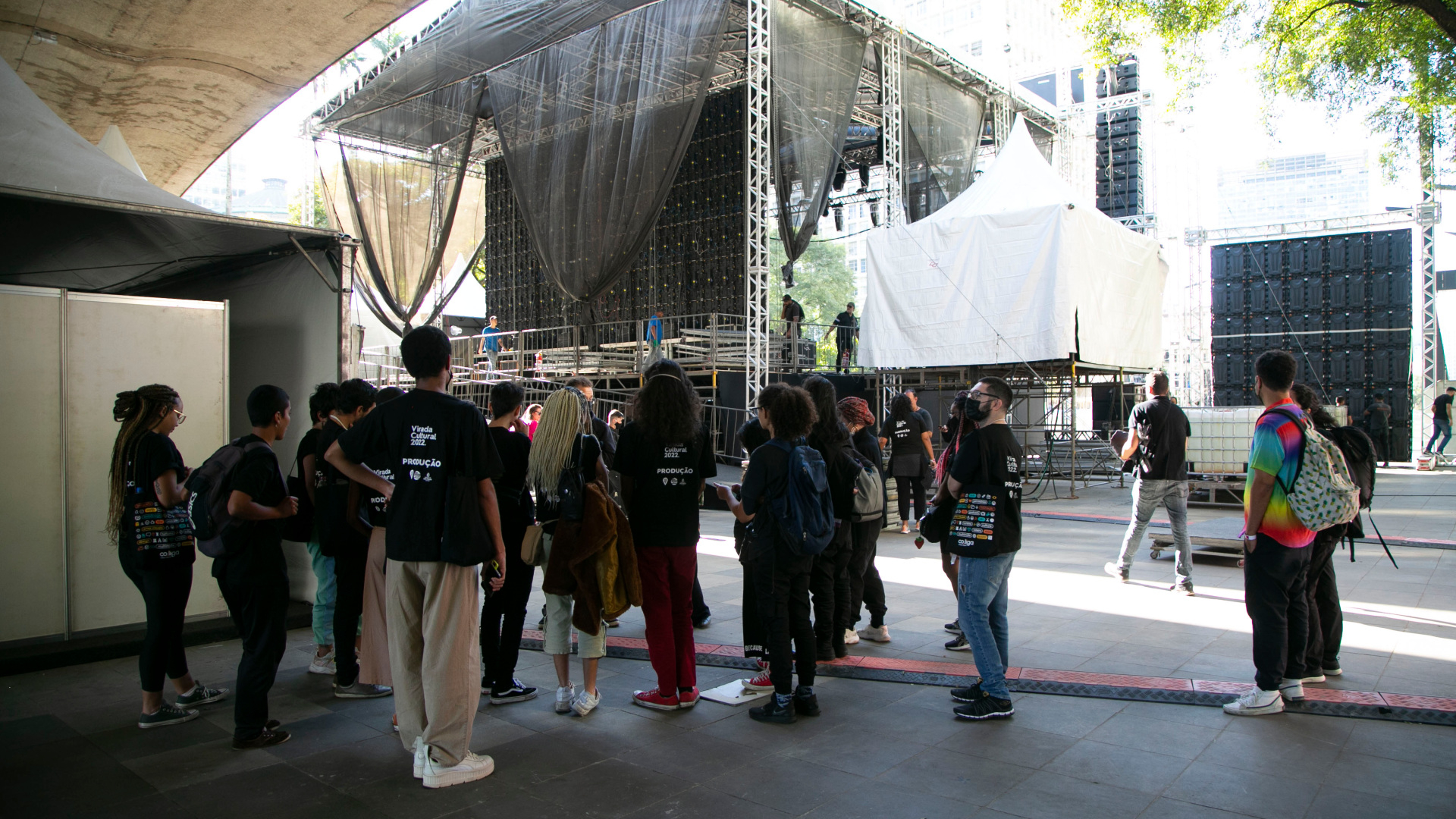 Pessoas estão reunidas, em pé, diante de um palco que está sendo montado.
