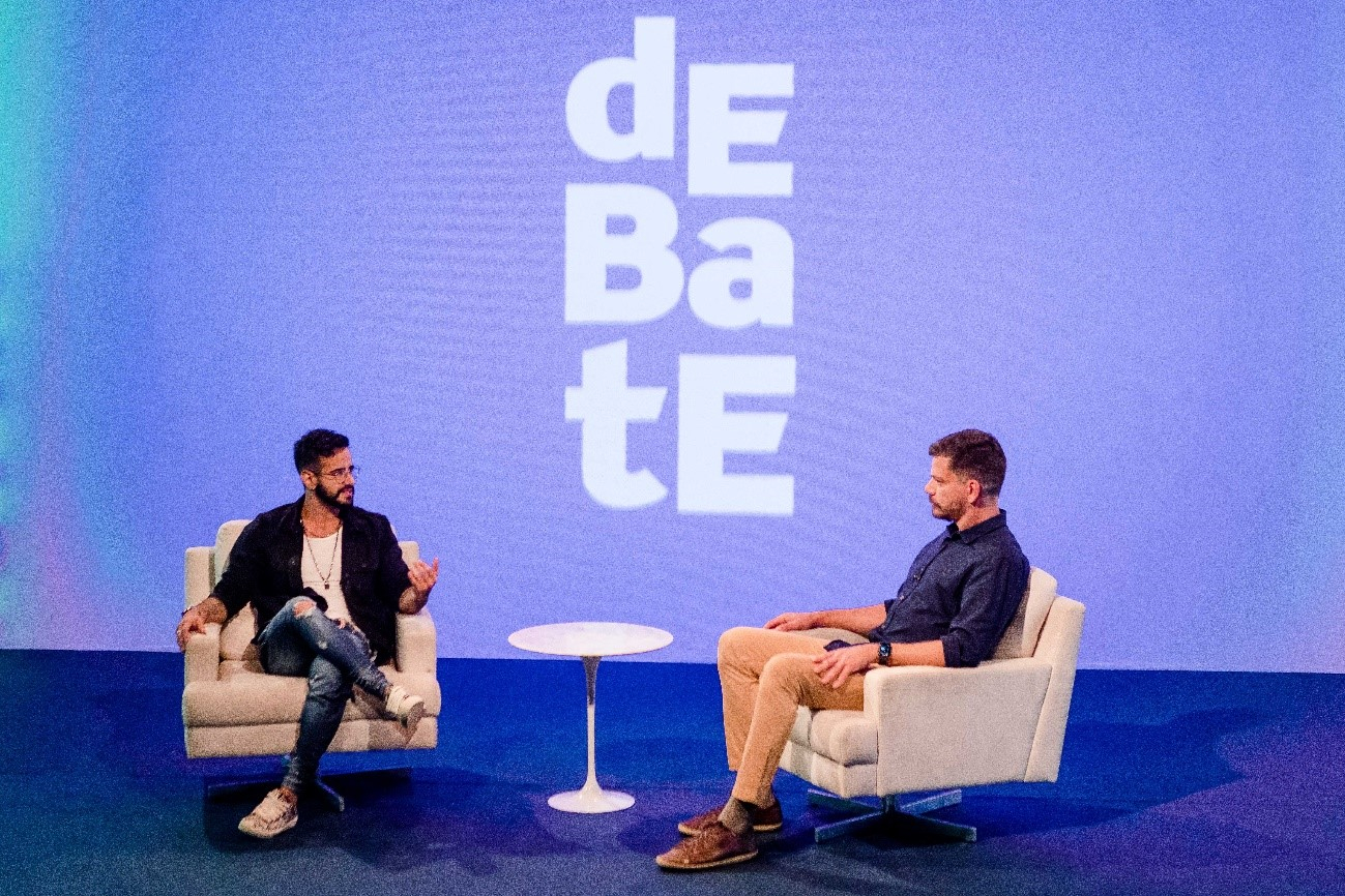Dois homens brancos sentados em poltronas brancas de frente um para o outro conversando. 