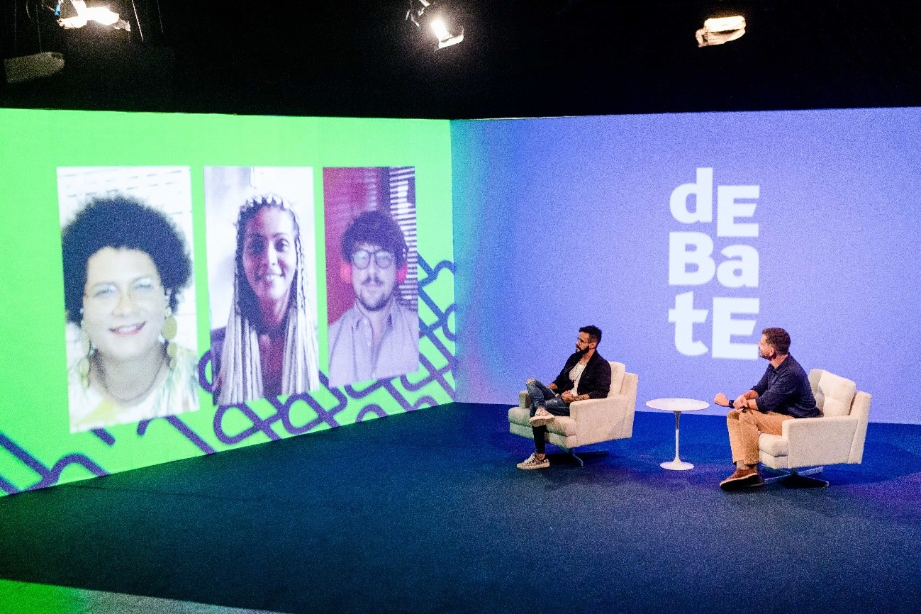 Um palco com dois homens brancos sentados em poltronas brancas e olhando para um telão com três pessoas.