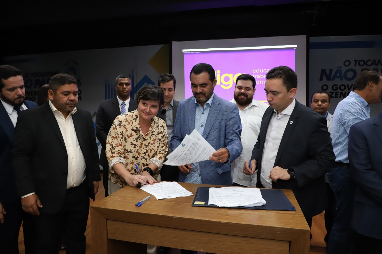 Uma mulher sorri enquanto participa de cerimônia de assinatura de contrato. Há diversos homens ao redor.