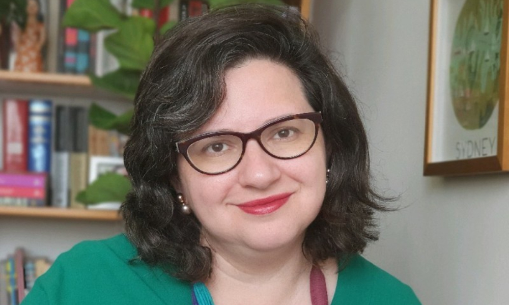 Uma mulher branca sorri. Ao fundo, uma estante com livros.