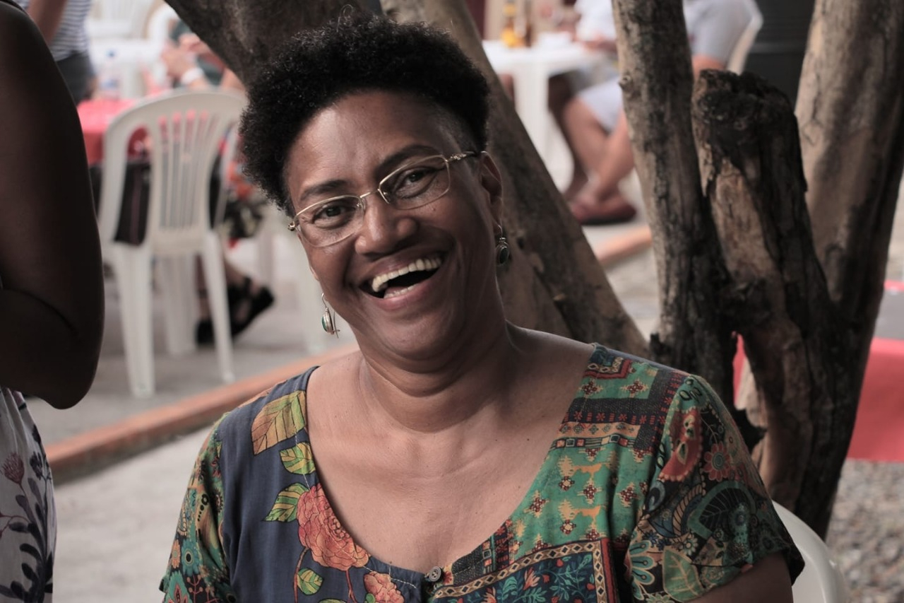 Uma mulher negra sorri. Ao fundo, troncos de uma árvore e, mais atrás, cadeiras de pvc brancas.