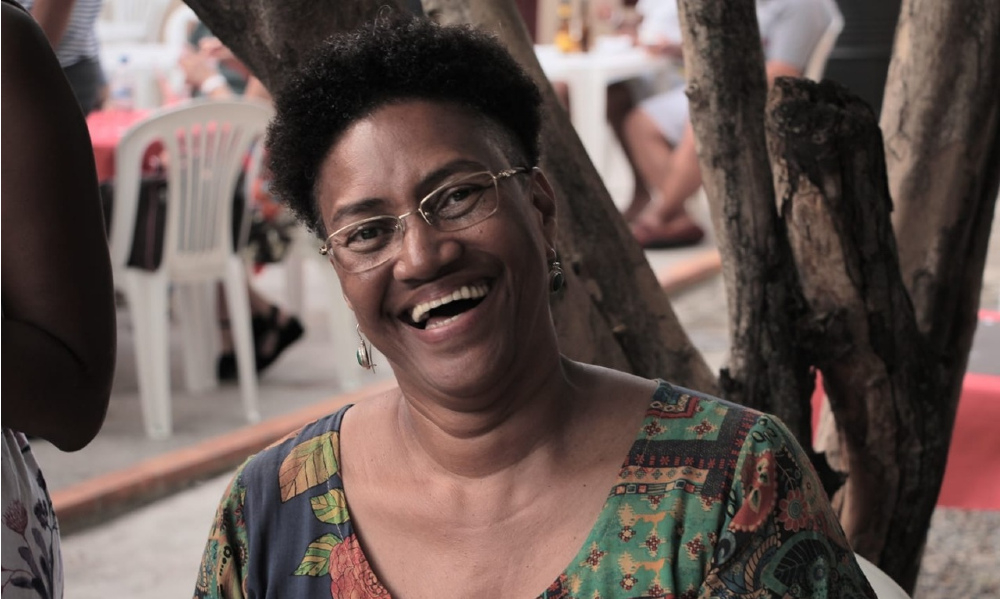 Uma mulher negra sorri. Ao fundo, uma árvore e algumas cadeiras de plástico.