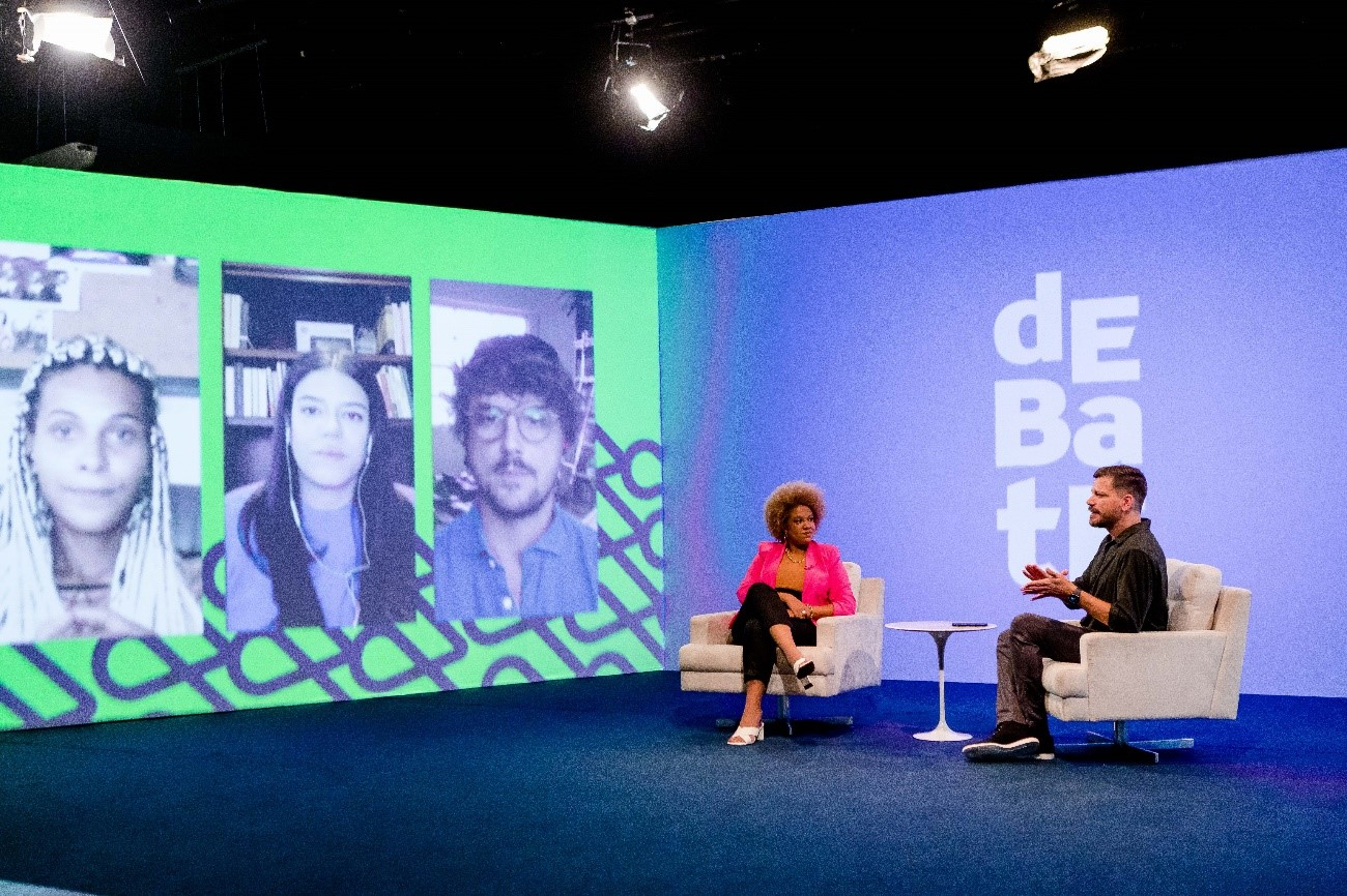 Uma mulher e um homem conversam, sentados em poltronas no estúdio, com 3 pessoas participando por videochamada da conversa.