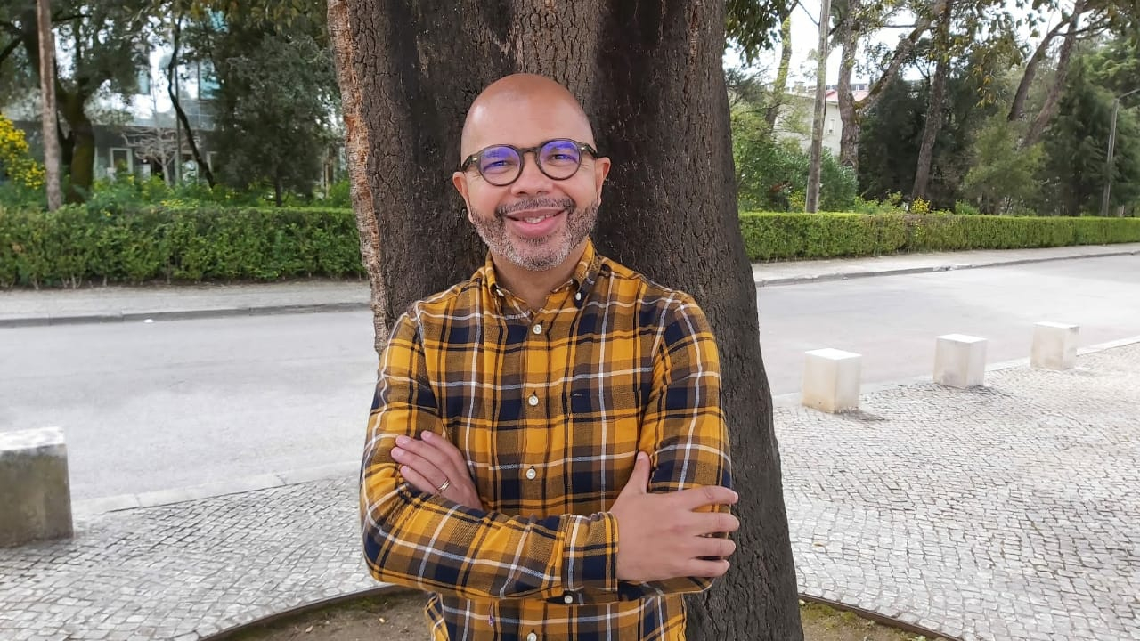 Um homem negro sorri. Ele usa camisa xadrez em tons amarelos e escuros. Atrás dele, o tronco de uma árvore.