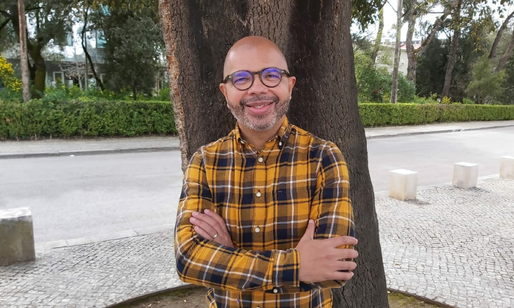 Um homem sorri. Ao fundo, uma árvore e uma rua.