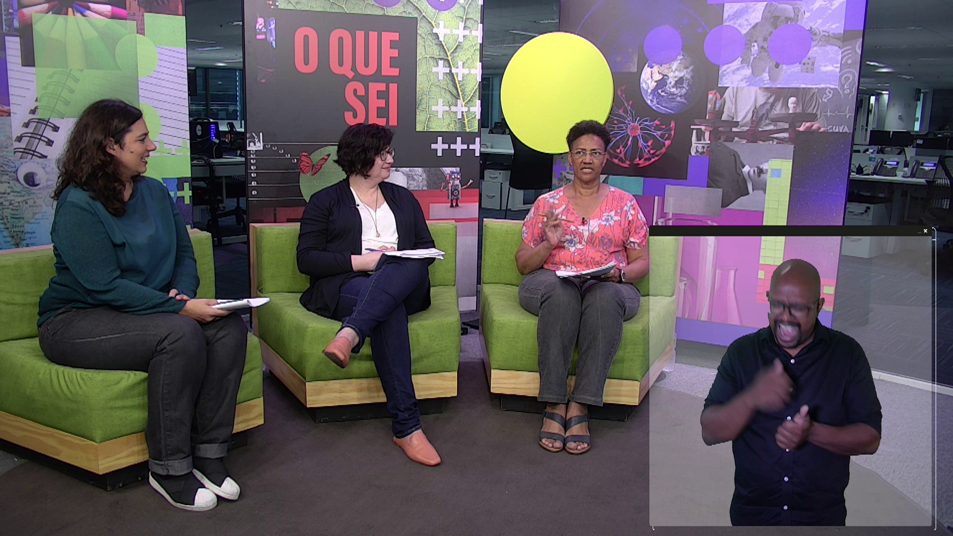 Três mulheres conversam em um estúdio de tv, sentadas em poltronas verdes. No canto inferior direito, o intérprete de Libras.