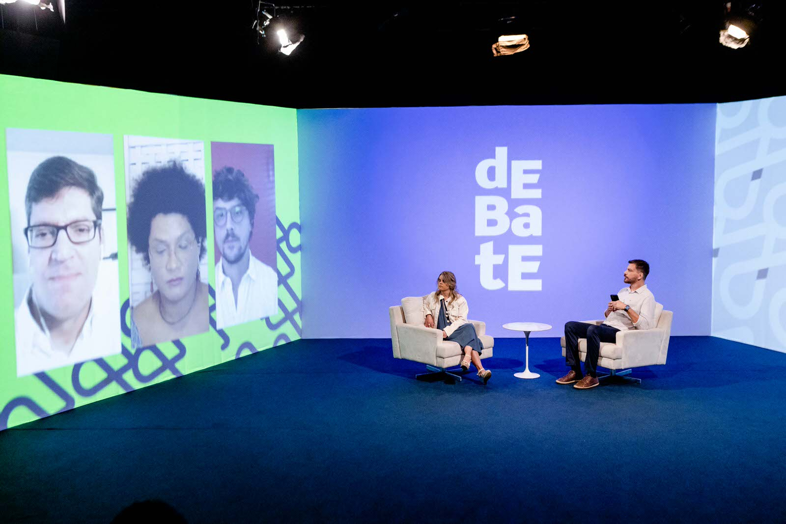 À esquerda, três pessoas têm suas imagens projetadas na parede do estúdio do programa. No centro e à esquerda do espaço, uma mulher e um homem estão sentados em poltronas, virados para as pessoas que estão projetadas na parede.
