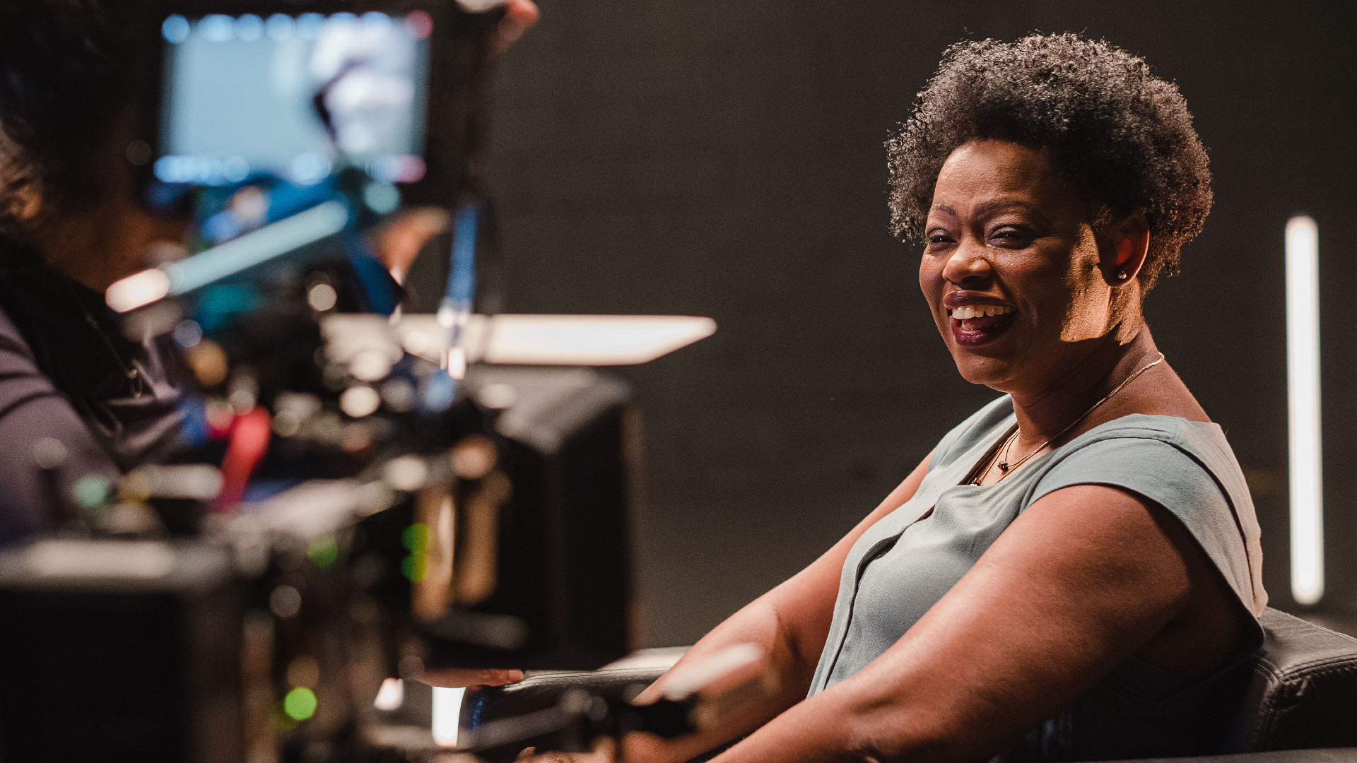 À direita da imagem, uma mulher negra sorri. À esquerda, aparelhos de um estúdio de tv e uma tela (ao fundo).