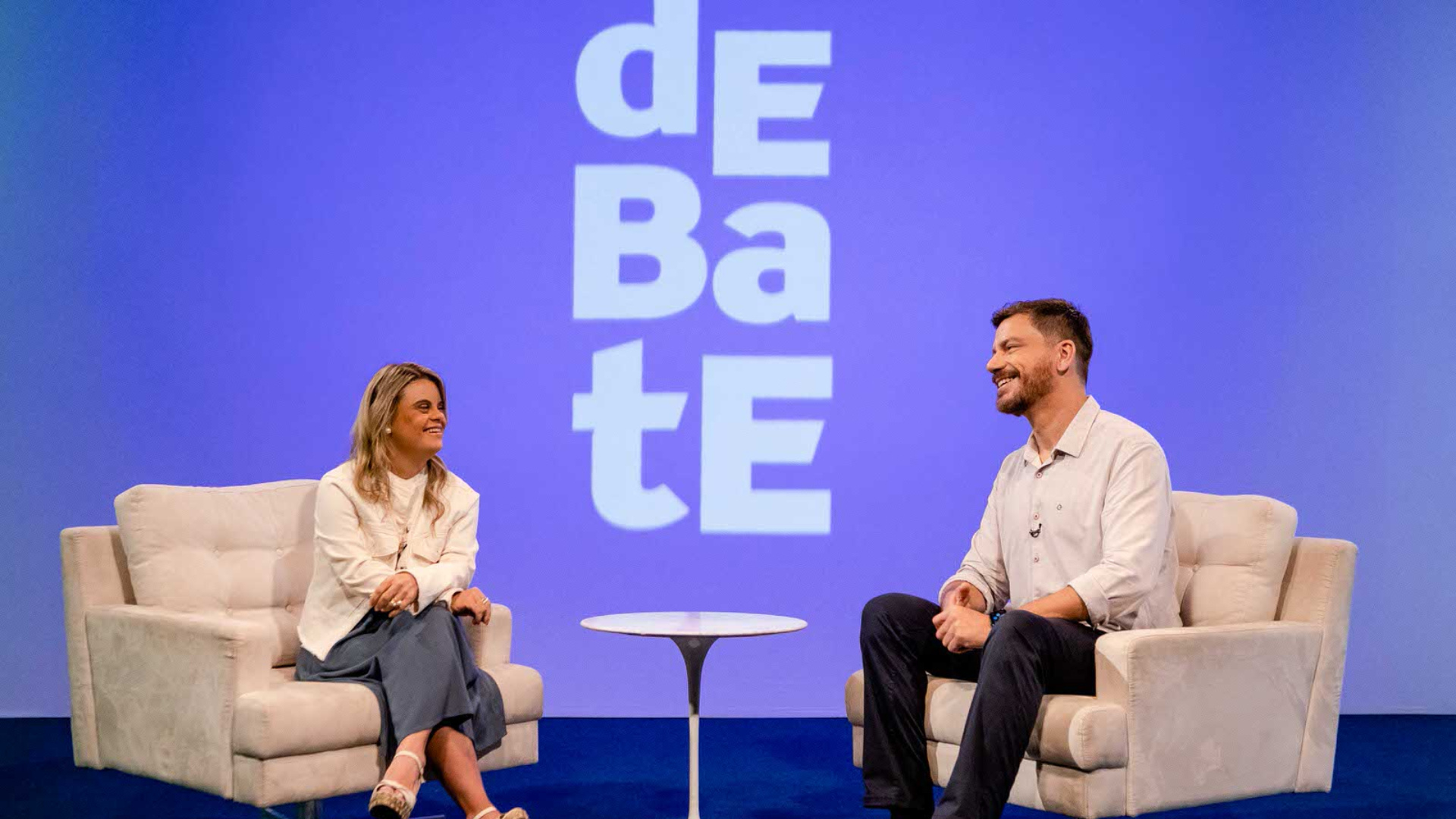 À esquerda, uma mulher branca, loira, com Síndrome de Down. À direita, um homem branco, de cabelo castanho. Ambos sentados em poltronas. A imagem tem o fundo azul com o nome do programa em tom mais claro (Debate).