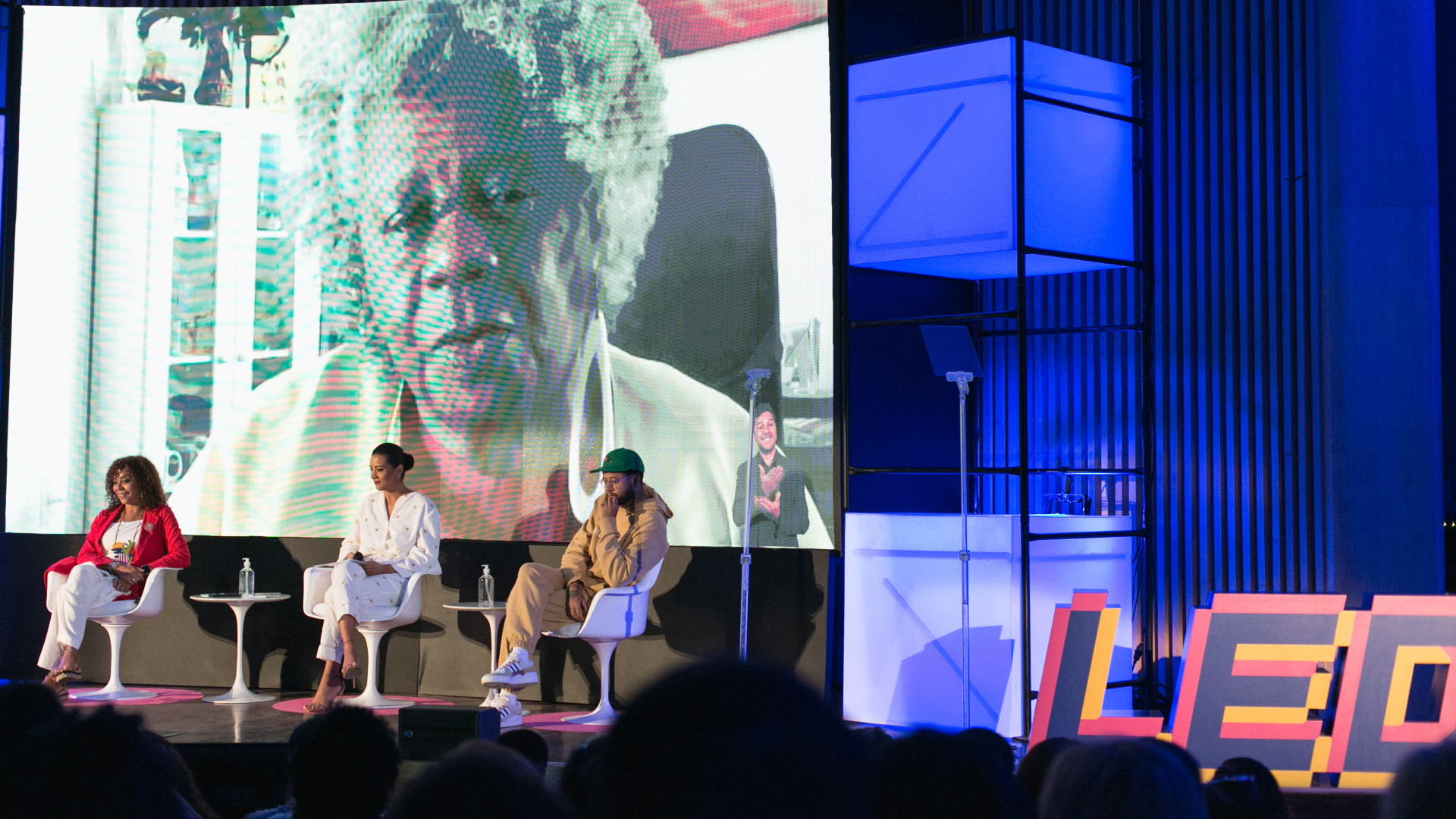 Três pessoas estão sentadas em poltronas, no palco de um auditório. Ao fundo, a imagem de Conceição Evaristo, mulher negra, com cabelo crespo e grisalho, é projetada em tamanho maior.