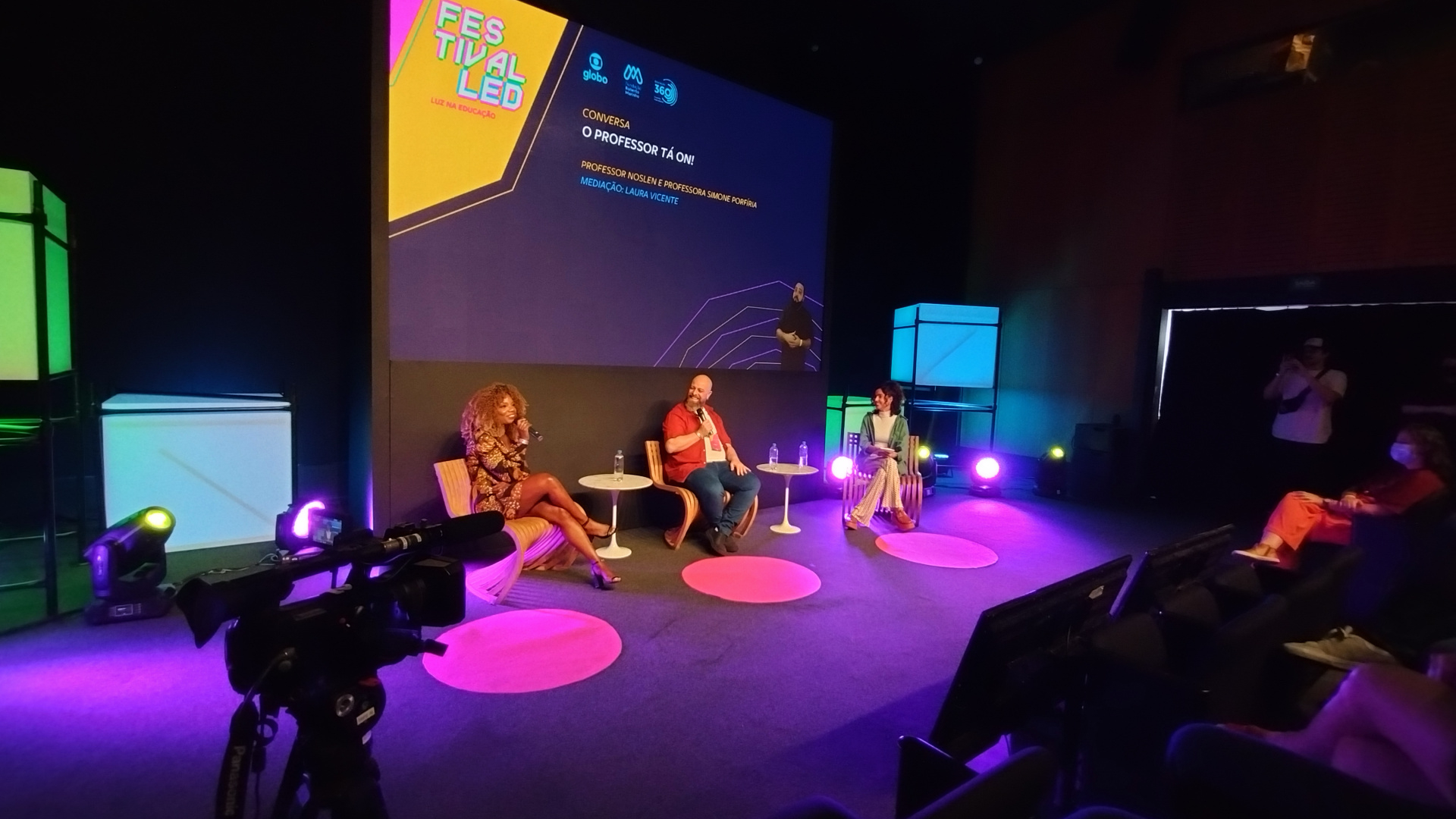 Três pessoas estão sentadas em poltronas em um palco roxo, com um telão ao fundo.