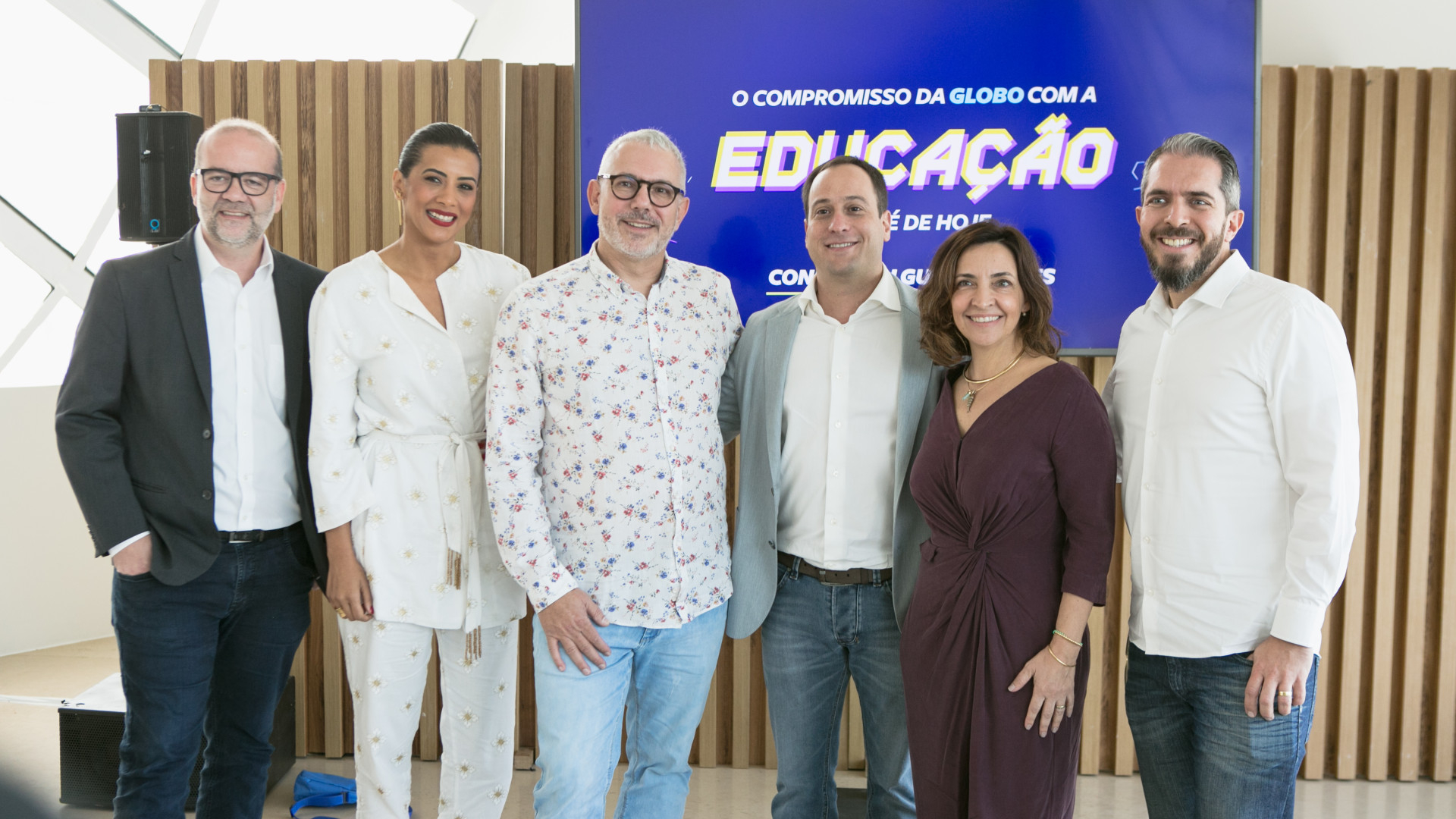 Seis pessoas estão em pé, sorrindo. Ao fundo, uma tela azul com o seguinte texto: "O compromisso da Globo com a educação".