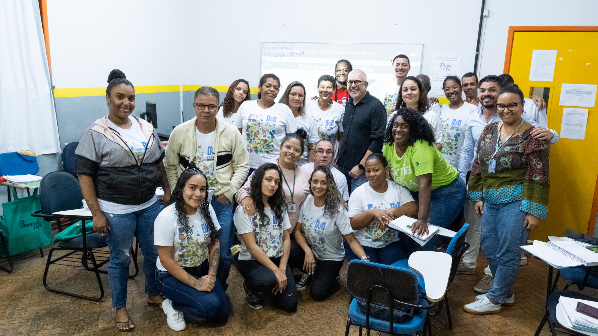 Diversas pessoas, em pé e agachadas, sorriem dentro de uma sala de aula.