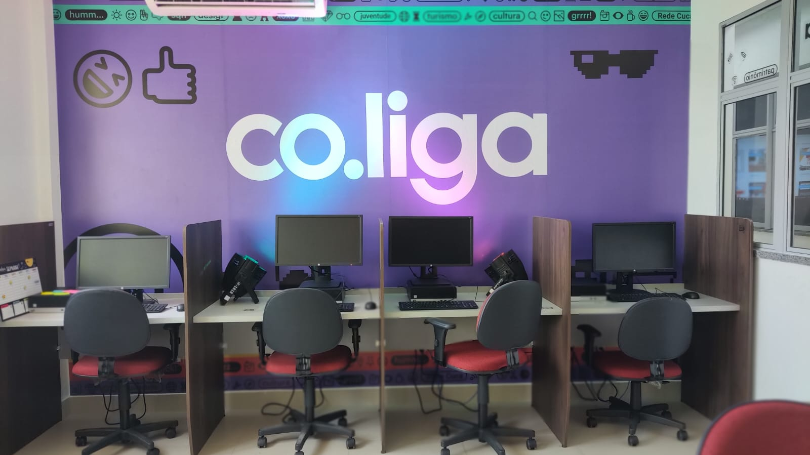 Uma sala de informática, com cadeiras diante de mesas com computadores. Ao fundo, a parede é lilás, com a logo da Co.liga.