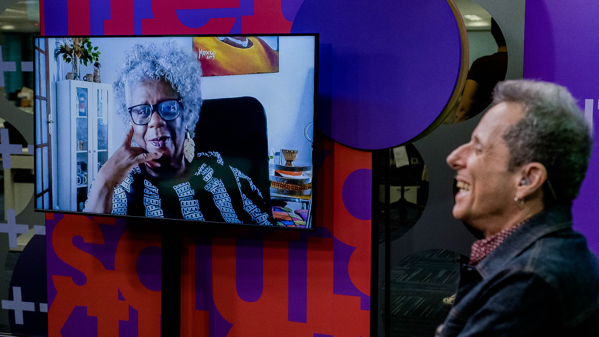 À esquerda, uma mulher negra, com cabelo grisalho, está na tela de uma tv, conversando com o apresentador do programa, que está à direita, sorrindo, de lado (mas de frente para a tv).