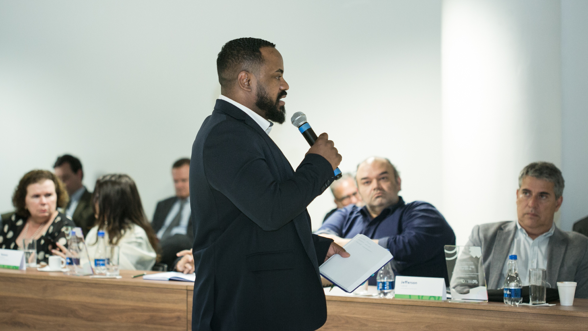 Homem negro fala, de pé, ao microfone, diante de uma plateia que o observa sentada.