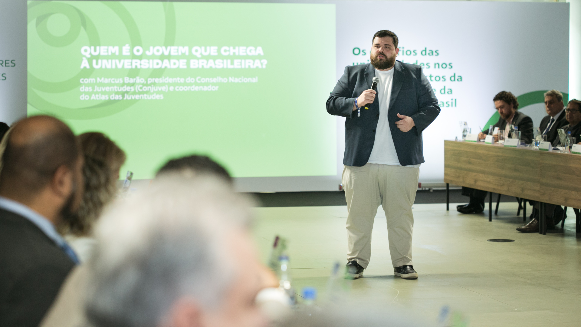 Um homem está de pé, falando ao microfone, , diante de uma plateia que o assiste, sentada, ao seu redor. 