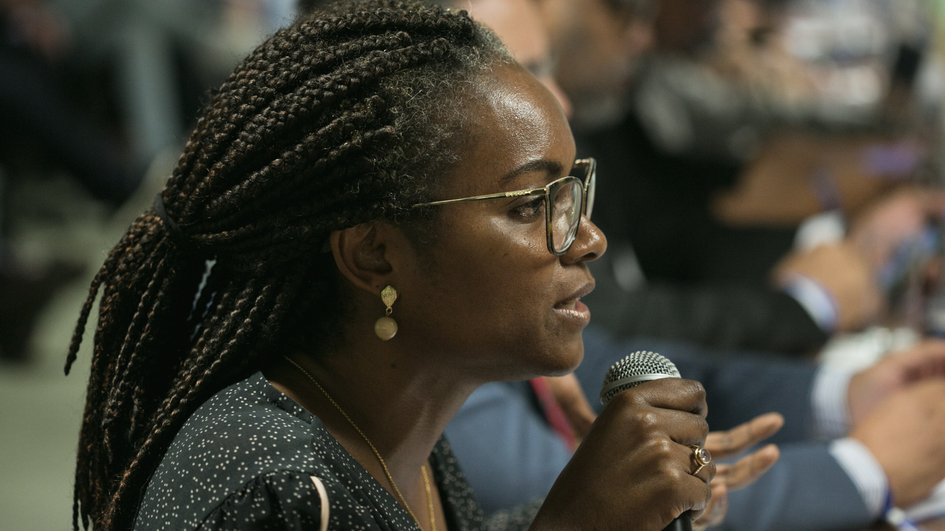 Uma mulher negra, que usa óculos e tem cabelos trançados em dreadlock, fala ao microfone.