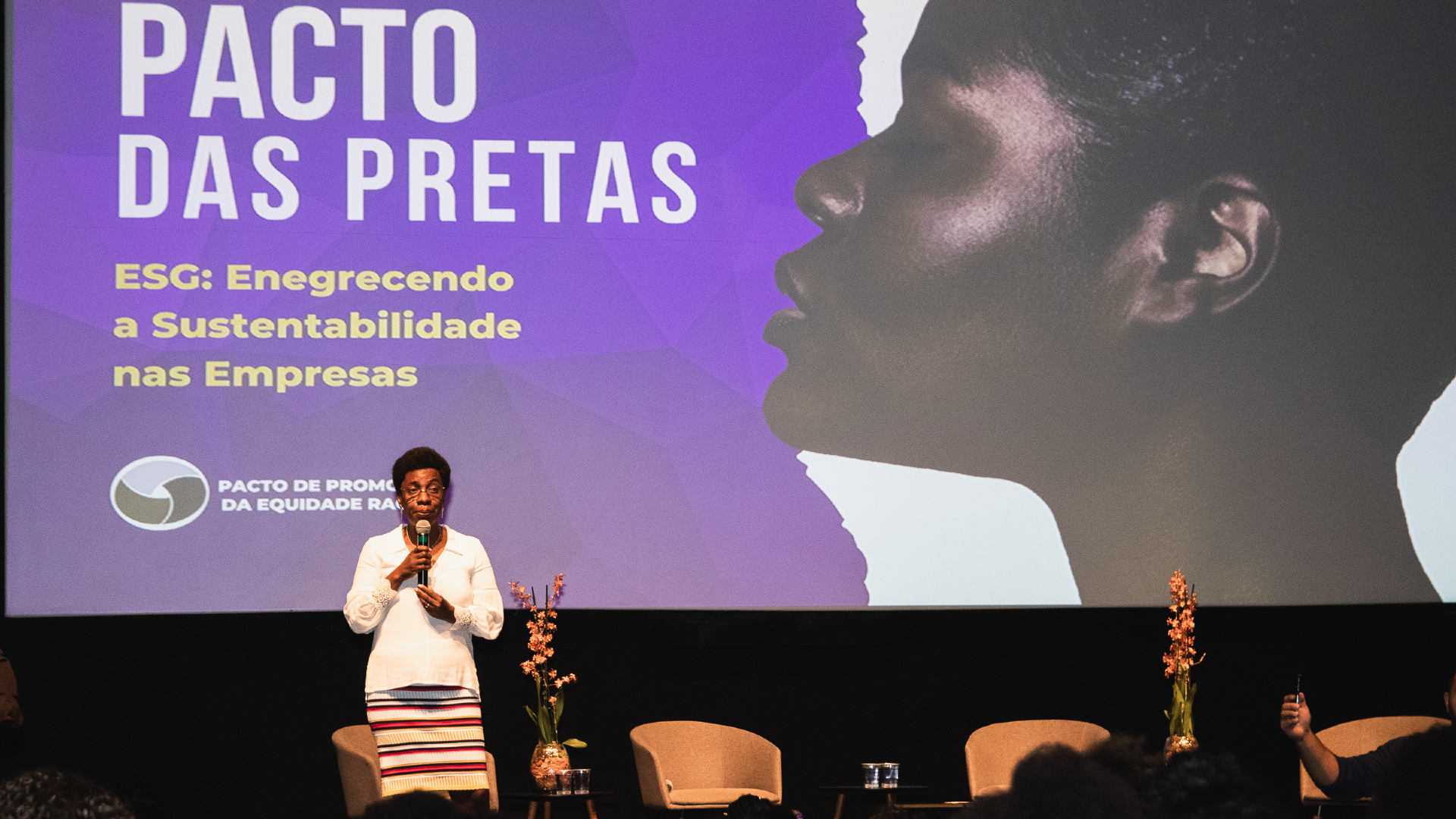 Mulher negra em pé no palco, usando uma camisa branca e saia listrada, diante do público. Ao fundo, o nome do fórum: "Pacto das pretas" e uma imagem de uma mulher negra de perfil.