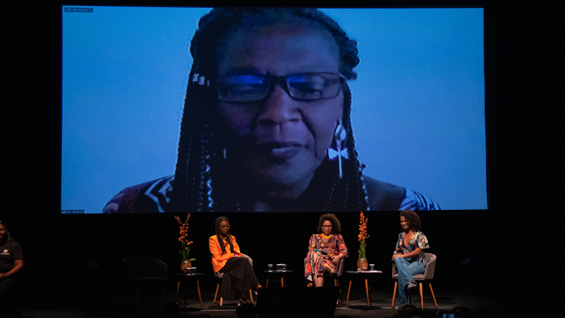 Três mulheres negras estão sentadas em cadeiras sobre um palco, diante do público. Em imagem projetada ao fundo delas, outra mulher negra. Esta, usando óculos.