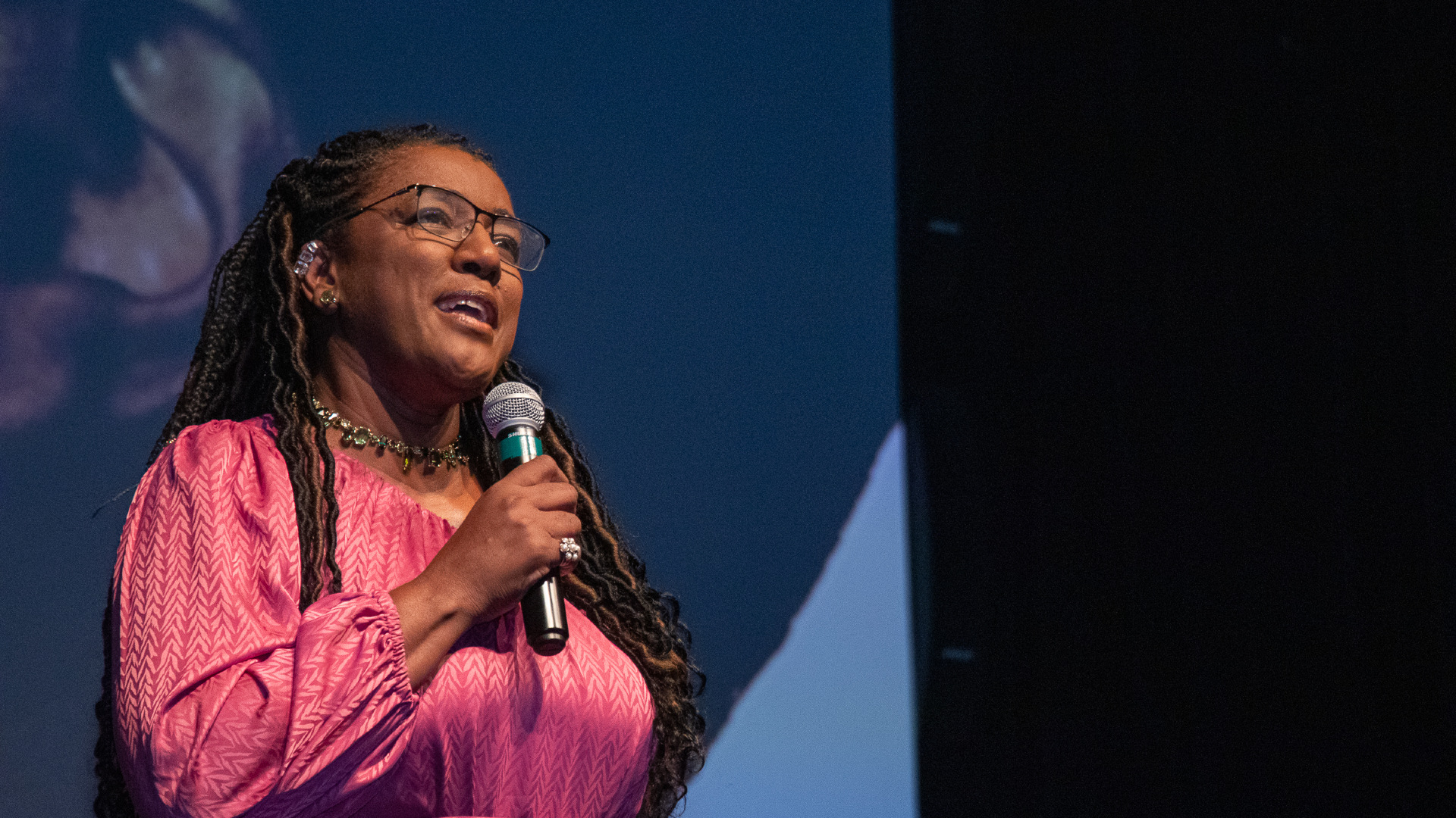 Mulher negra discursa com um microfone na mão, usando uma camisa lilás e dando um leve sorriso.