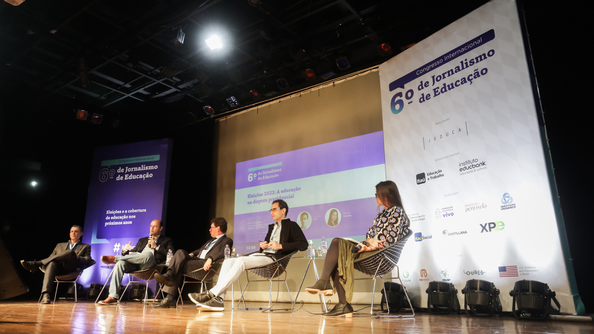 Cinco pessoas estão sentadas, lado a lado, em poltronas sobre um palco de madeira brilhosa.