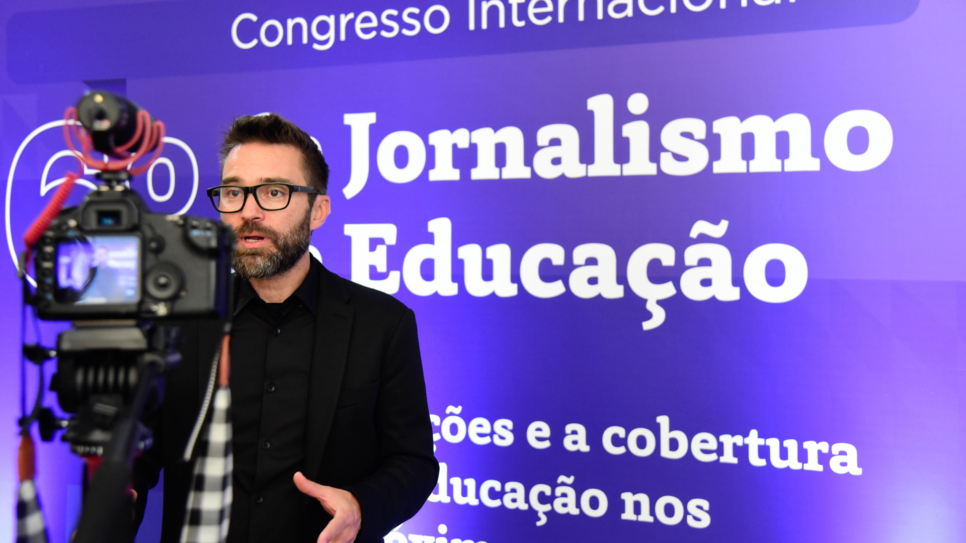 Homem fala diante de uma câmera. Ao fundo, em tela de cor de azul, as palavras "Jornalismo" e "Educação".