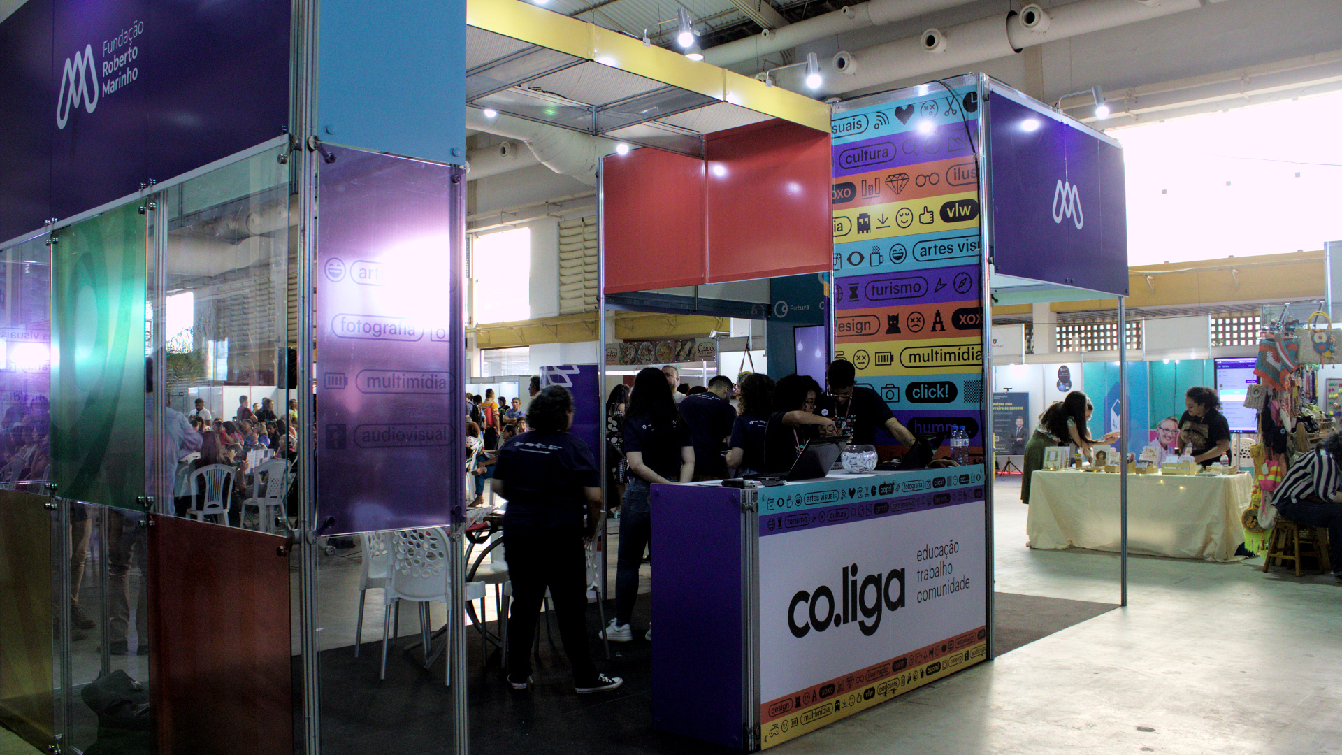 Estande com placas coloridas. Em destaque, a palavra "co.liga", que é a escola de economia criativa da Fundação Roberto Marinho.