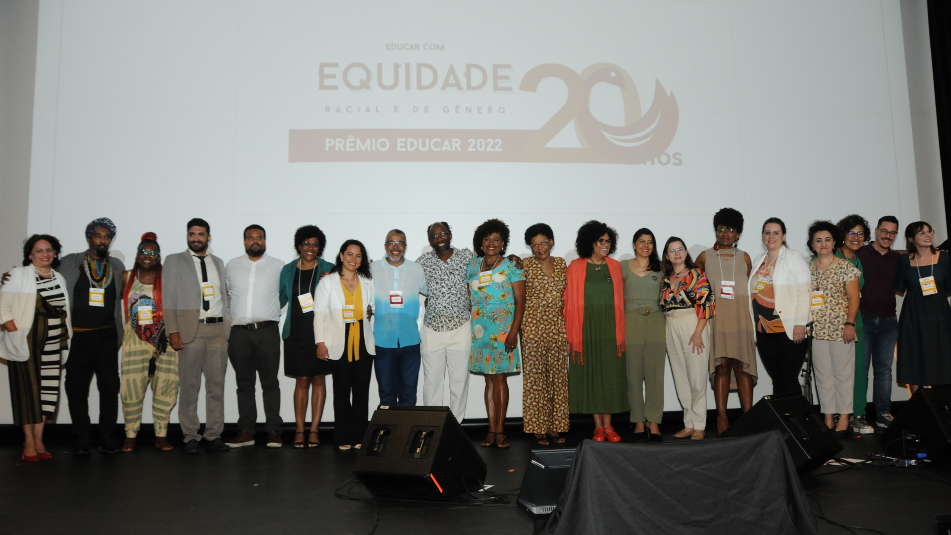 Diversas pessoas de pé, sobre um palco. Ao fundo, uma tela branca.