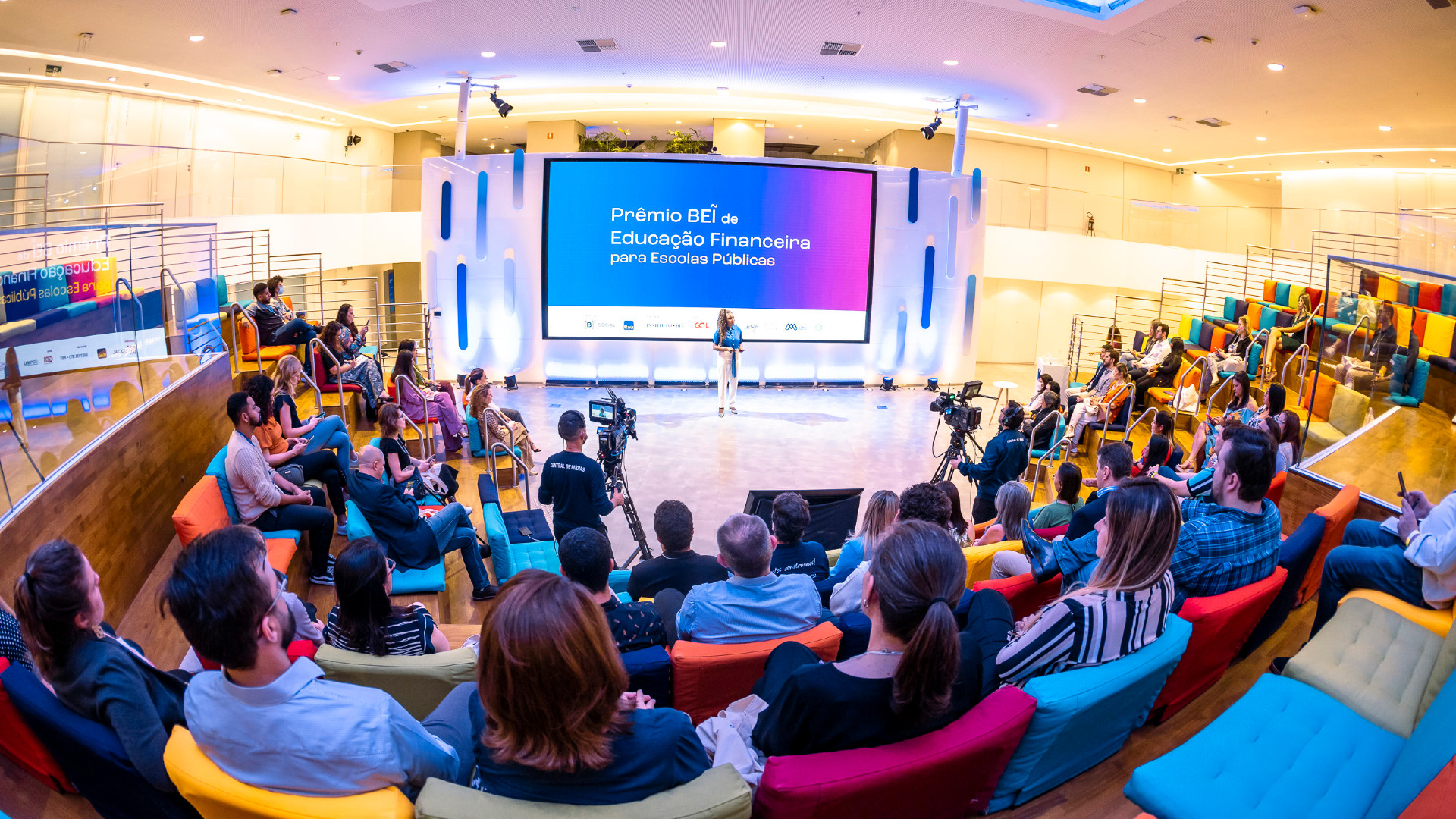 Várias pessoas sentadas em um auditório assistem a uma mulher que está no palco, ao fundo.