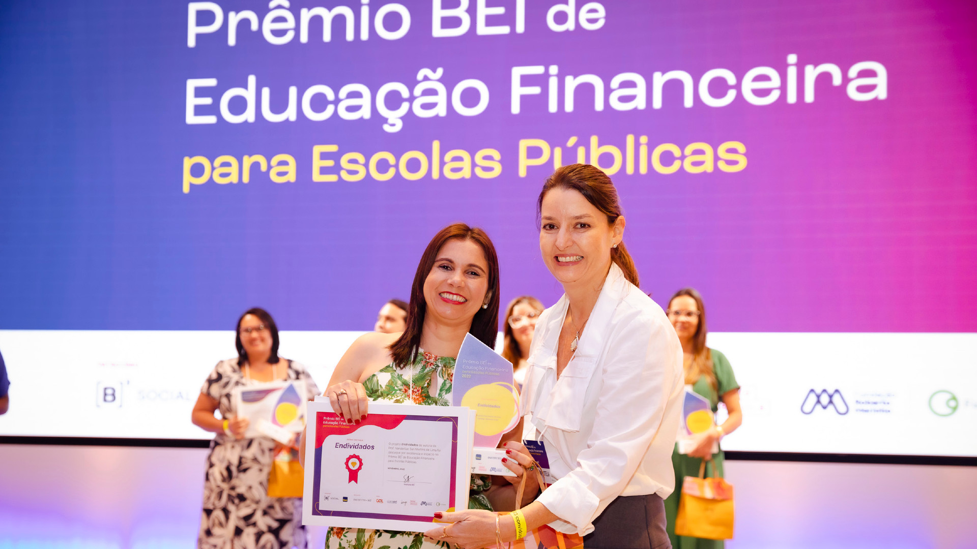 Uma mulher entrega um certificado de premiação a outra mulher. Ambas sorriem.