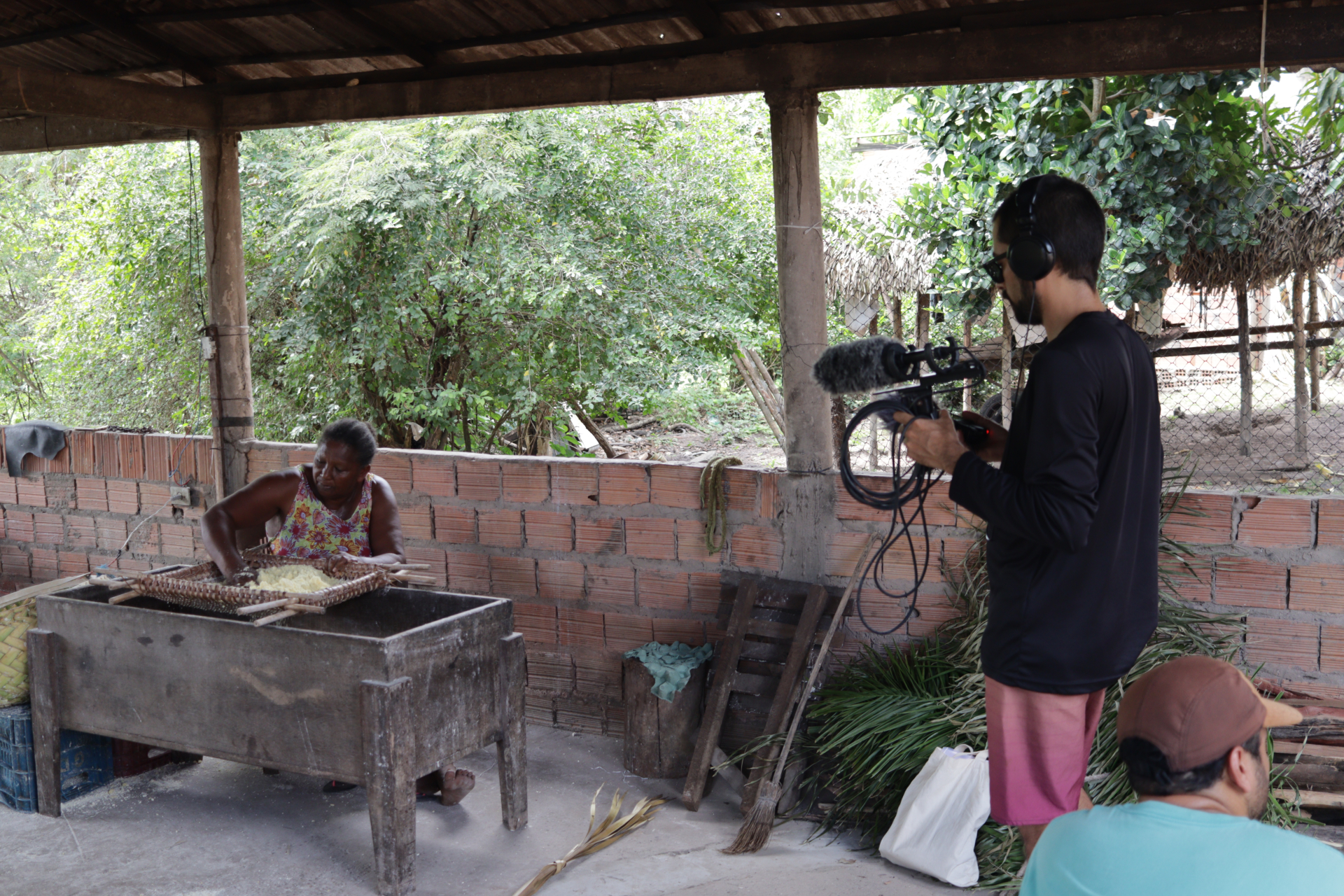 À esquerda, uma senhora negra faz um trabalho manual enquanto um homem a filma (este, à direita).