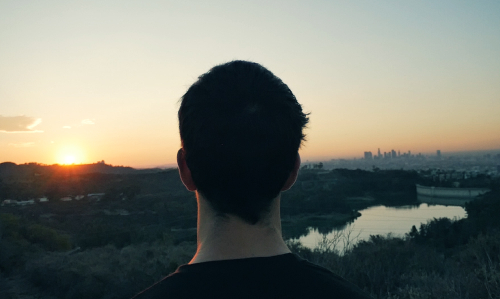 Em primeiro plano, de costas, um homem olha para o horizonte. O sol está nascendo ou se pondo.