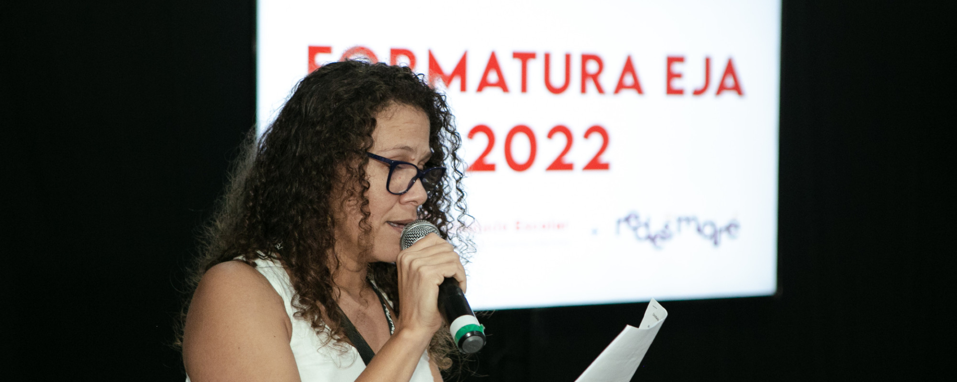 Mulher de cabelos enrolados, usando óculos, faz discurso que lê em um papel. Ao fundo, o texto projetado na parede: "Formatura EJA 2022".