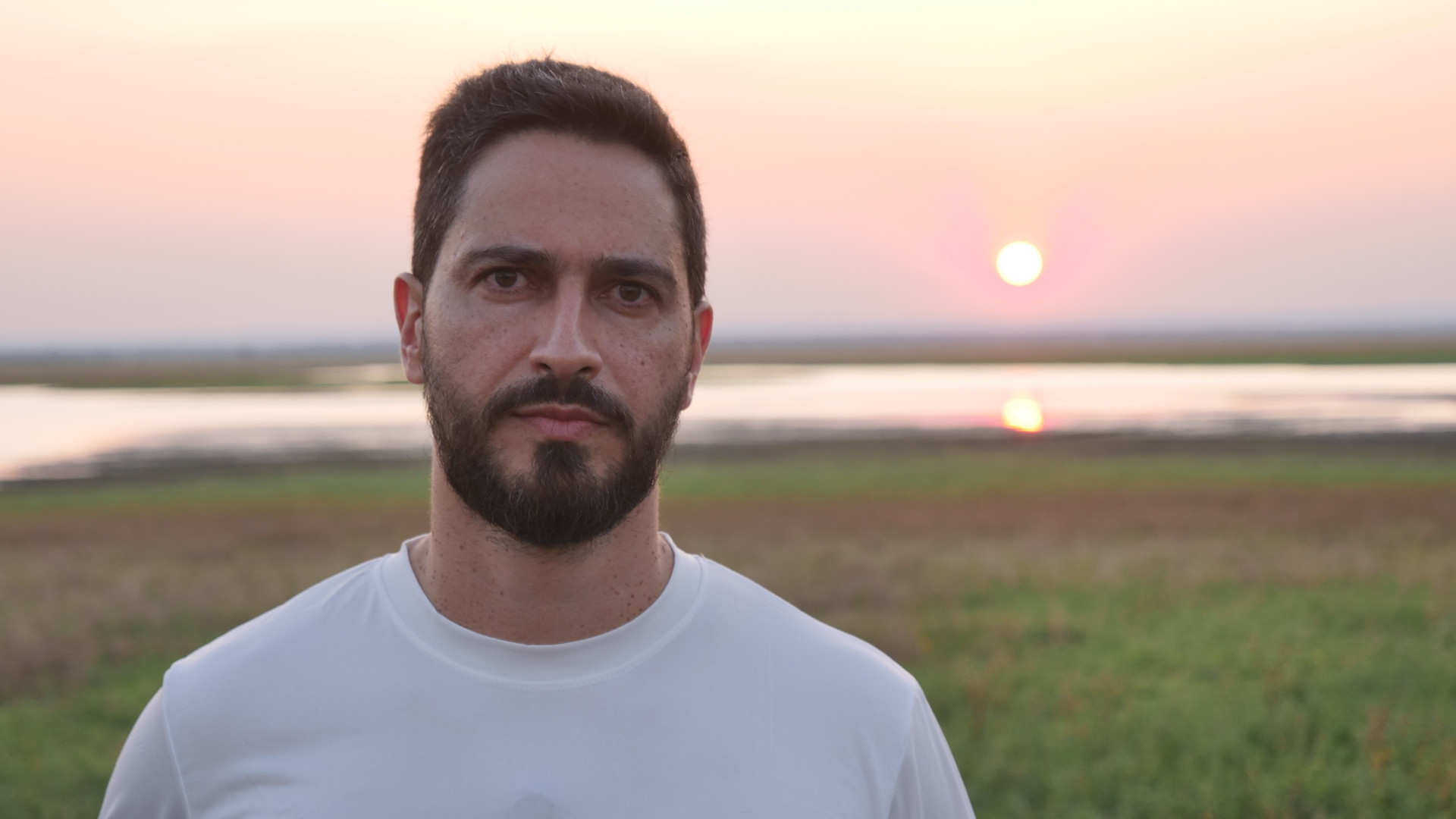 Homem branco, com cabelo e barba pretos, em primeiro plano. Ao fundo, o horizonte com o sol se pondo.