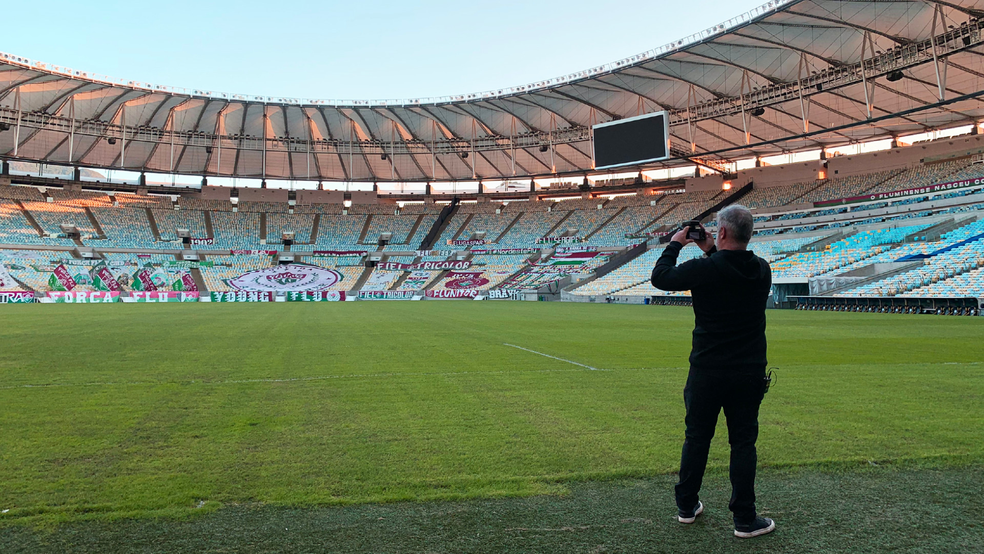 Homem está de pé no gramado de um estádio (e de costas para quem o fotografou) e parece estar fotografando a arquibancada que está ao seu redor.