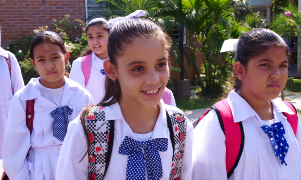 Quatro meninas estão vestidas com o que parecem ser uniformes escolares. Uma delas sorri.